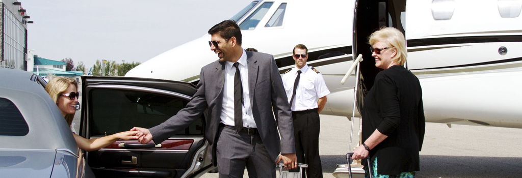 Fully vetted and trusted chauffeur in a suit and tie helping a smiling, professional woman out of a car with private jet and trusted pilot in the background.