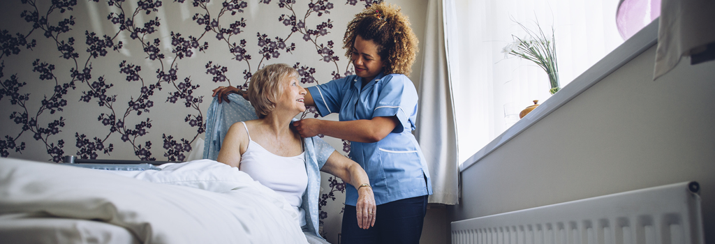 Trusting, female patient is smiling at a young, female, african-american nurse who is assisting her dress in a nursing home setting.